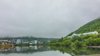 A foggy morning in the city. The houses are reflected in the mirror-smooth water of the lake. Green vegetation on the shore. The mountains are hidden in a thick mist. Petropavlovsk-Kamchatsky. 