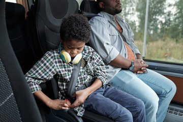 Serious Afro American boy with headphones around neck sitting in bus chair and buckling up before...