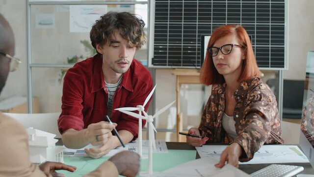 Multiethnic team of engineer sitting at office table with models of windmill turbines on it and discussing papers while working on project of alternative energy