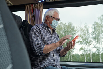Aged Caucasian man in mask and eyeglasses sitting at window and using phone while sending sms in bus