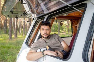 Naklejka premium Young bearded man with book looking at camera in window of mobile house in the forest