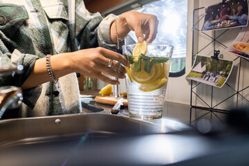 Hands of young woman putting sliced lemon and mint leaves into jug with water while making lemonade