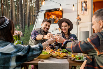 Group of young cheerful friends toasting with wine by served table against travel house