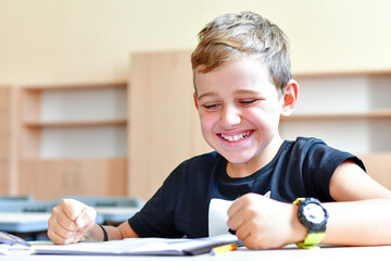 Happy smiling  schoolboy  cute clever schoolboy  in glasses sitting alone  at desk at empty classroom