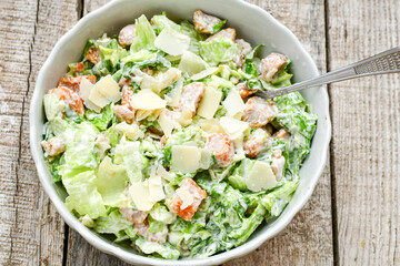 Homemade traditional Caesar Salad with Parmesan Cheese and Croutons in a white bowl on a old white table with leaves and a sauce boat 