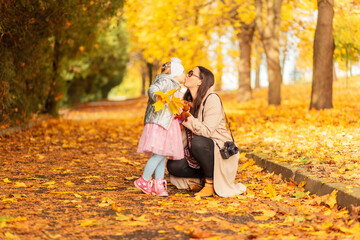 Fototapeta premium Mom and daughter in fashionable clothes walk in the park and make a kiss on the background of yellow autumn foliage