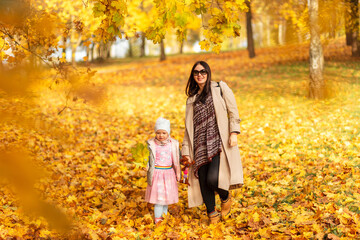 Happy beautiful mother woman with her daughter child in fashionable clothes are walking in the autumn park with yellow foliage outdoors
