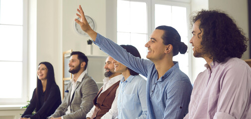 Happy young man in the audience raises his hand to ask a speaker a question. Banner with a group of...