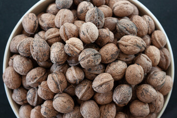 Closeup of a bunch of self-harvested homegrown whole unpeeled healthy walnuts with shell stacked in a white bowl used for decoration and food snack on a black granite countertop background in top view