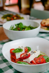 Caprese salad with strawberry, mozzarells, basil and balsamic sauce served in a white bowl over green plaid background.