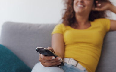 Relaxed woman sitting on the couch at home and watching TV, she is holding the remote control