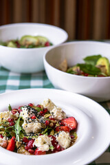 Green salad with cucumber, zucchini and strawberries in a white bowl over green plaid tablecloth.