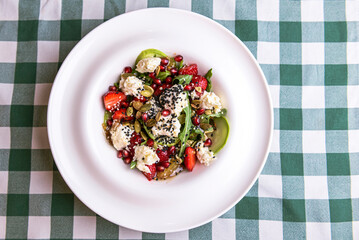 Green salad with cucumber, zucchini and strawberries in a white bowl over green plaid tablecloth.