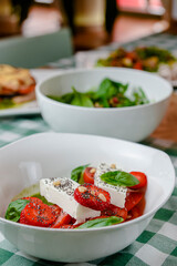 Caprese salad with strawberry, mozzarells, basil and balsamic sauce served in a white bowl over green plaid background.