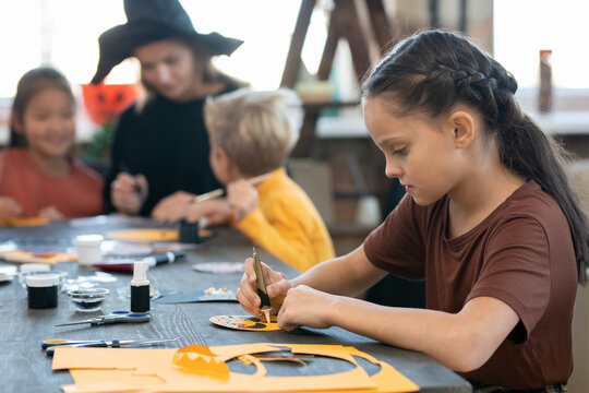 Side view ofo little girl with oil paint preparing halloween paper symbols by table