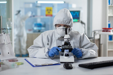 Biologist researcher wearing ppe medical suit analyzing genetics sample using clinical microscope in microbiology hospital laboratory. Scientist woman working at vaccine development expertise