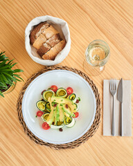 from above fresh raw zucchini salad with tomatoes and avocado accompanied by a glass of white wine and a cloth basket with bread