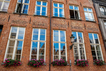 Fototapeta na wymiar Blossoming flower pots under the windows of a beautiful old brick house, street view of old Bruges, Belgium, cloudy summer day with window reflections