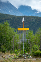 A disc golf hole basket on the ground with the woods and mountains in background during summer time in Norway.