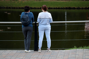 Two women standing near the metal railing on embankment on the pond, leisure in autumn park 