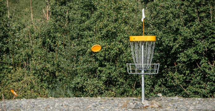 A disc flying towards the "hole" in the disc golf basket which is on the ground with the woods in background during summer time in Norway.