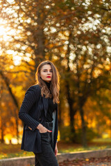 Fototapeta na wymiar Stylish beautiful young woman in fashionable elegant clothes with a black blazer, sweater and jeans walking in a park with bright yellow autumn foliage at sunset