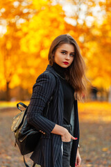 Fototapeta na wymiar Beautiful young girl with red lips in a fashionable blazer with a black sweater, chain and backpack walks in nature with golden autumn foliage at sunset