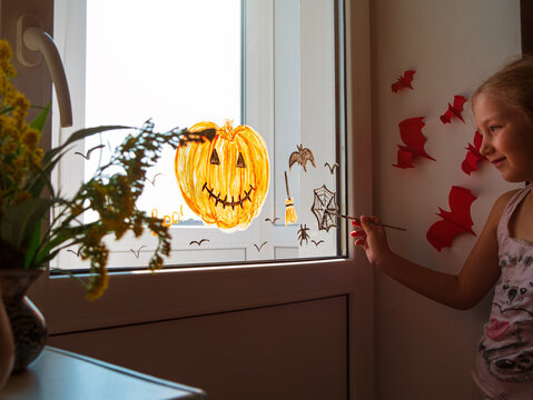 Child painting pumpkin on window preparing to celebrate Halloween. Little girl decorates her room autumn holiday at home