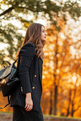 Fototapeta na wymiar pretty elegant woman in fashionable black suit with stylish blazer and leather backpack walks in the park with colored yellow autumn foliage at sunset