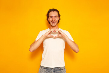 happy cheerful smiling man in casual clothes folded his hands in the shape of a heart in front of his chest. Isolated on yellow background with space for text. concept of people, love, relationships