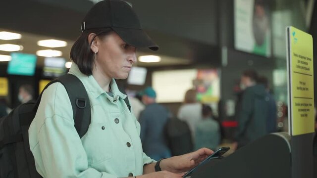 A woman in a baseball cap near the terminal at the airport scans the QR code from the smartphone screen. Self check-in terminal.