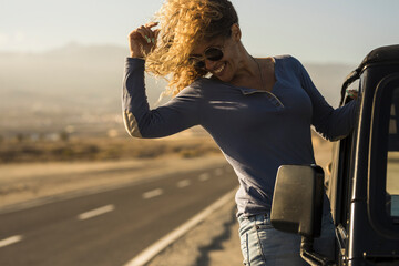 Fototapeta premium Cheerful woman in sunglasses hanging on side door of car at roadside. Young woman enjoying vacation on road trip. Carefree woman having fun leaning on outside of car door on her vacation trip