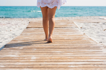 Low section of woman walking on wooden plank amidst sand leading towards sea water. Woman walking towards sea through wooden planked footpath along the sandy beach - Powered by Adobe