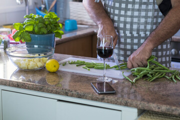 Man cutting fresh beans on cutting board with knife in kitchen. Raw vegetables for dinner preparation on kitchen platform. Male chef in apron cutting fresh vegetables to cook food in domestic kitchen