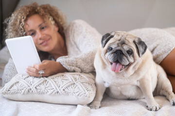 Young woman with her pet pug using digital tablet. Cute pet pug dog with female owner lying on...