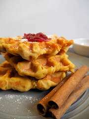 on a gray plate are gluten-free pumpkin Belgian waffles, sprinkled with powdered sugar and red currant jam, with cinnamon sticks on a gray background . side view