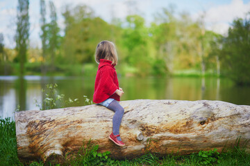 Adorable preschooler girl sitting on log and having fun in spring forest or park