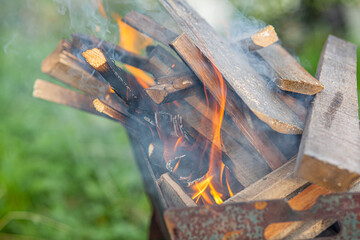The firewood in the grill burns with a bright orange flame of fire on a natural green background. Preparation for cooking meat on the grill in nature. Fire flames and smoke 