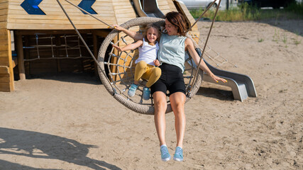 Mom and daughter swing on a round swing. Caucasian woman and little girl have fun on the playground.