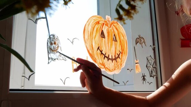 Child painting pumpkin on window preparing celebrate Halloween. Little kid draws decorates room interior with paper bats celebration autumn holiday at home Creative family leisure lockdown new reality