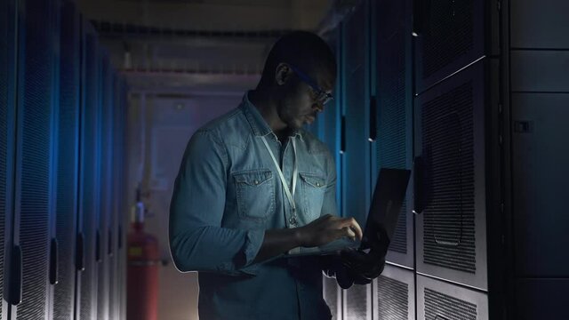 Young male engineer working with laptop hardware while standing in render farm spbas. Close-up view of American African man holds computer in hand and looks at screen, checks equipment and stands in