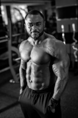 Fototapeta na wymiar Black and white photo of the strong man with muscular body type posing in modern sports hall. Multiracial man looking at the camera with light smile