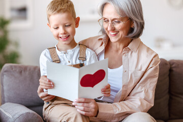 Cute little boy grandson congratulating smiling grandmother and giving handmade greeting card