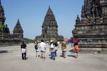 Group of Asian people with tour guide walking around ancient hindu Prambanan Temple complex. Sunny clear blue sky background.