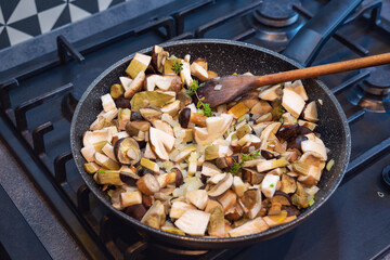 Frying pan full of chopped, fresh bay boletes and boletus, decorated with green twig during the process of frying. Gas stove in the background.
