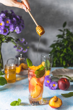 Woman hand perfused honey on peach infused water, cocktail, lemonade or tea. Summer iced cold drink with peach and mint on light gray table surface