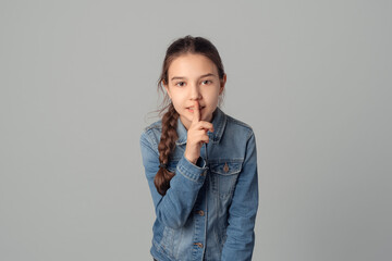 Young girl holds finger to his mouth, shows TSS, SHH, asks to remain silent about her mistake, isolated on grey background, wearing in denim jacket