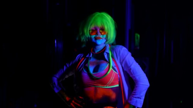 a woman with green hair and red glasses is dancing on a dark background. the skin is painted with bright colors. the camera is moving