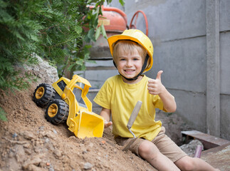 cheerful 5-year-old boy in yellow T-shirt and in a construction helmet plays with toy bulldozer at...