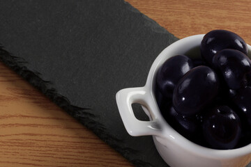 Composición en la que aparece una tapa de aceitunas sobre un plato de pizarra en una mesa de madera